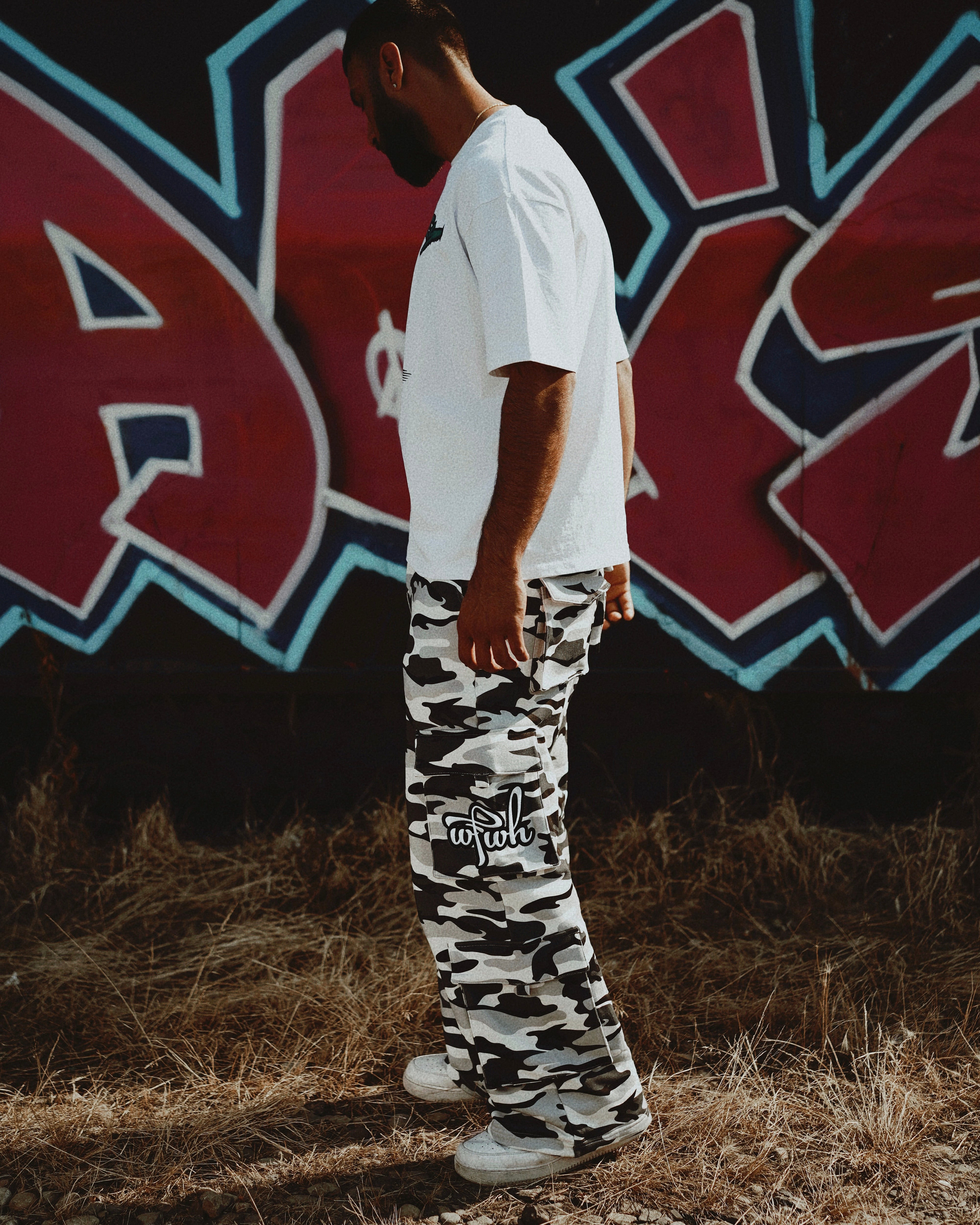Person wearing a white hip hop t-shirt and wfwh camouflage pants standing in front of a graffiti wall.