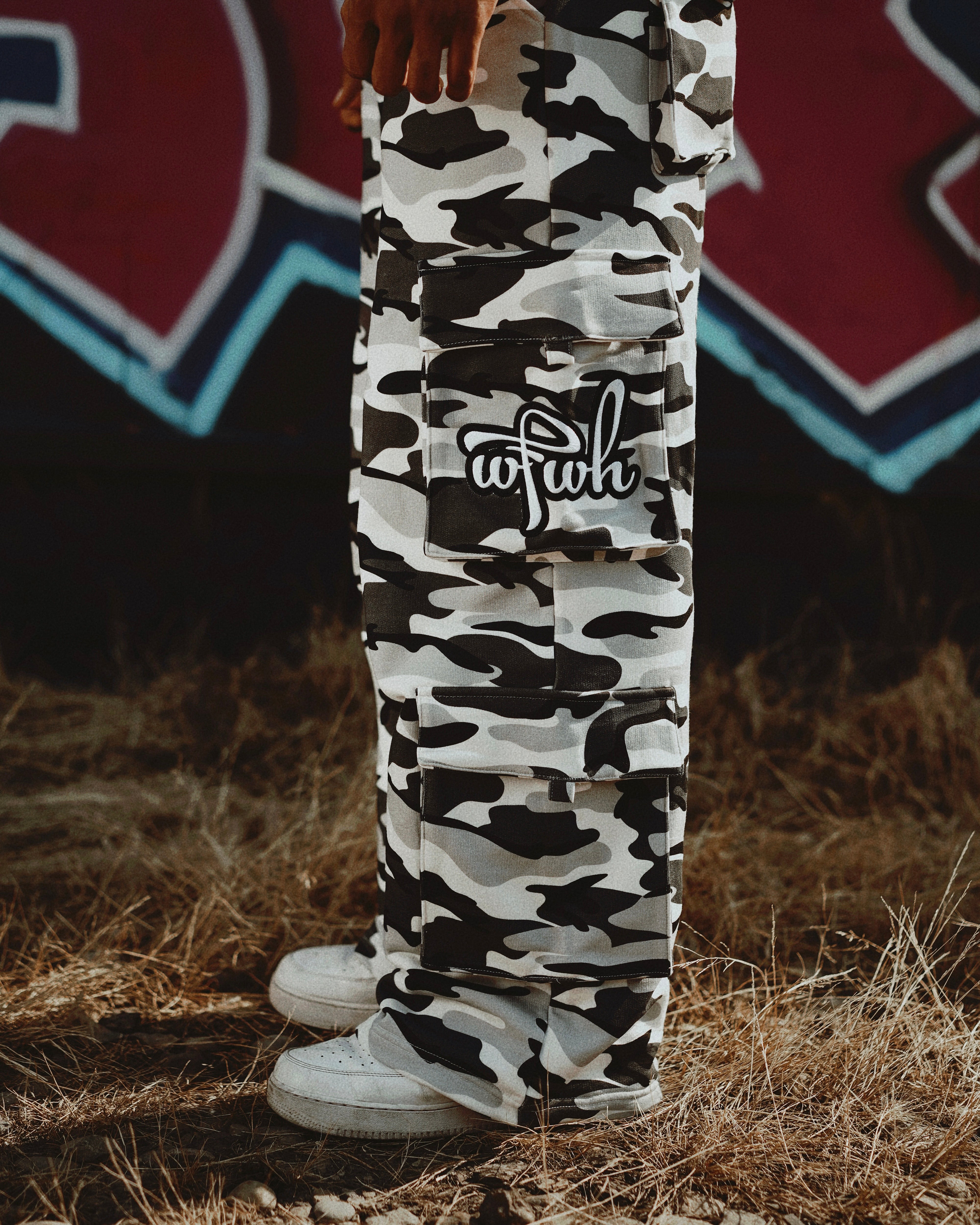 Person wearing wfwh camo pants with a logo, standing on dry grass with a blurred background